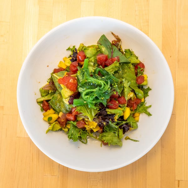 A vibrant salad with green lettuce, red tuna, yellow mango, and green seaweed in a white bowl.