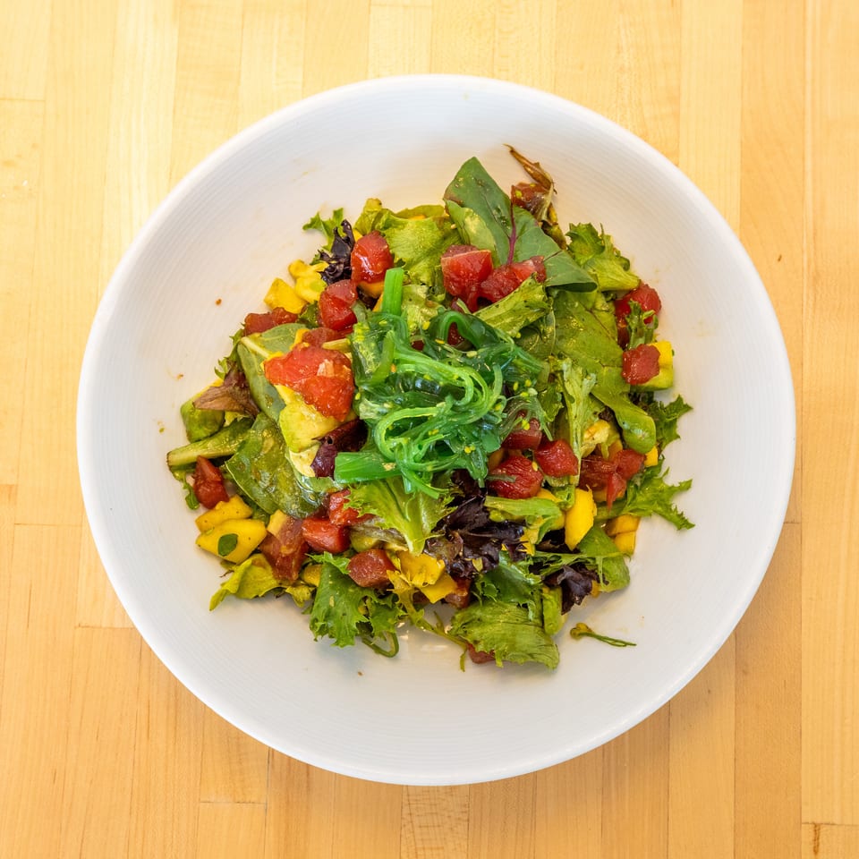 A vibrant salad with green lettuce, red tuna, yellow mango, and green seaweed in a white bowl.