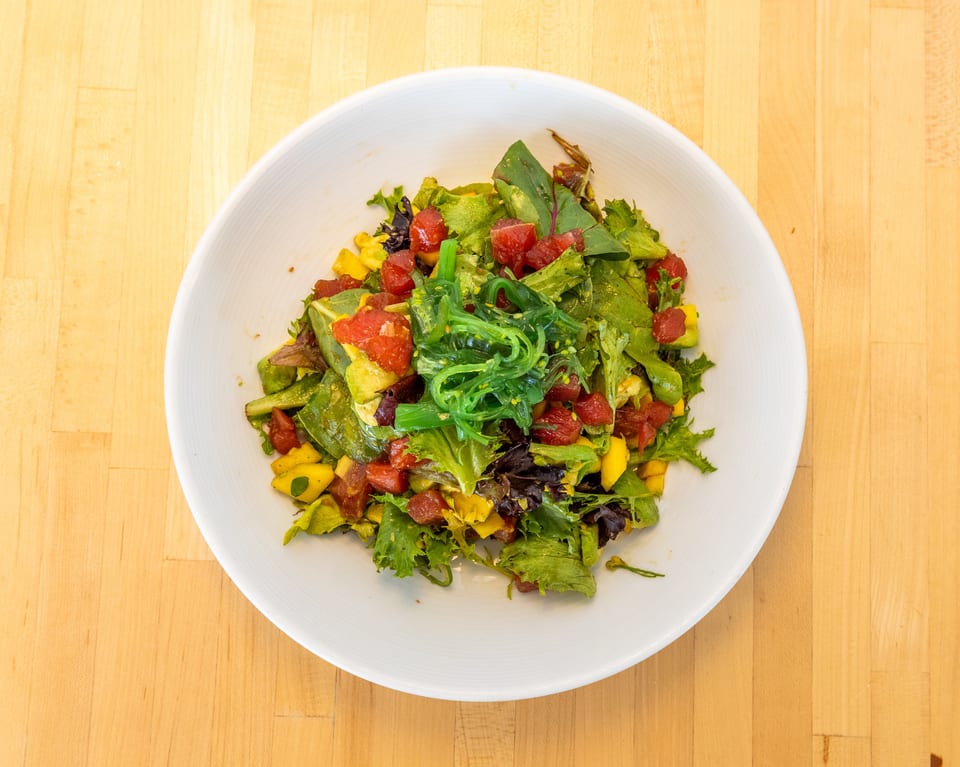 A vibrant salad with green lettuce, red tuna, yellow mango, and green seaweed in a white bowl.