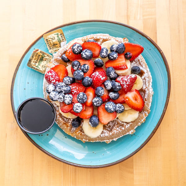 Waffle topped with strawberries, blueberries, bananas, powdered sugar, syrup, and butter on a blue plate.