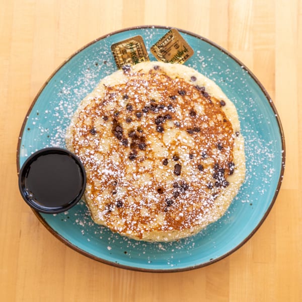 Chocolate chip pancake with powdered sugar, syrup, and butter pats on a blue plate.