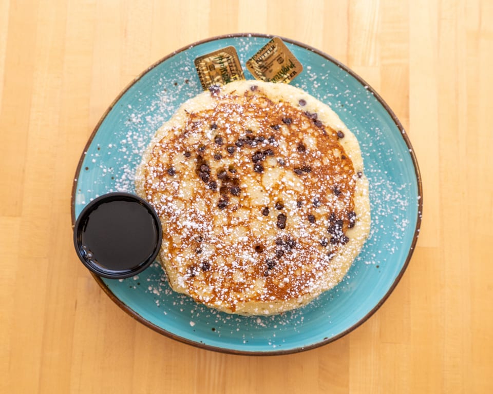 Chocolate chip pancake with powdered sugar, syrup, and butter pats on a blue plate.