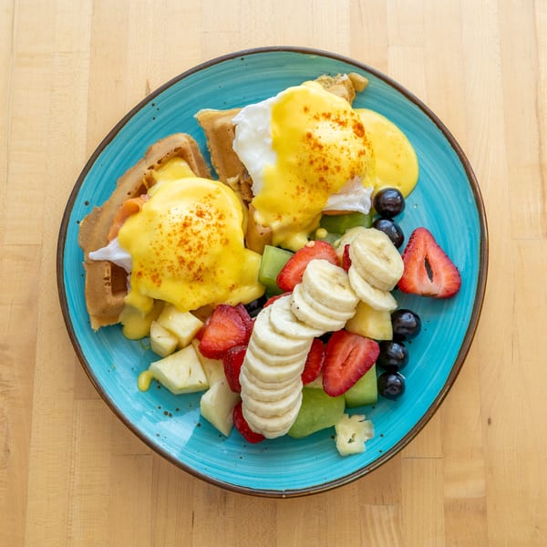 Eggs Benedict on waffles with hollandaise sauce, served with a side of fresh fruit.