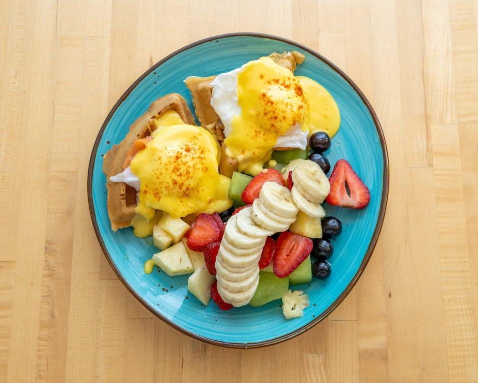 Eggs Benedict on waffles with hollandaise sauce, served with a side of fresh fruit.