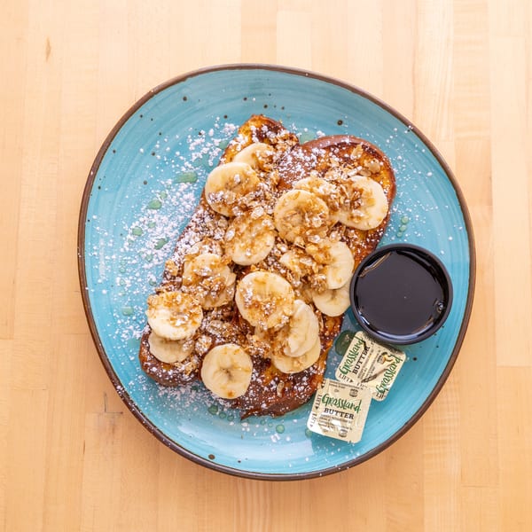 French toast topped with sliced bananas, granola, powdered sugar, and syrup on a blue plate.
