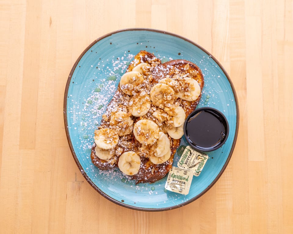 French toast topped with sliced bananas, granola, powdered sugar, and syrup on a blue plate.