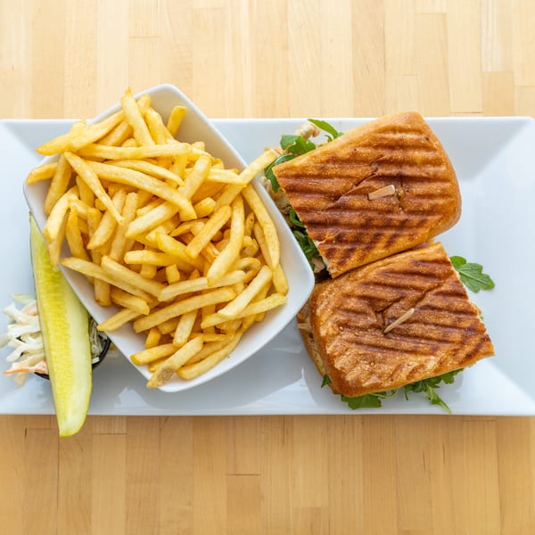 Grilled panini sandwich with fries, coleslaw, and a pickle spear on a white rectangular plate.