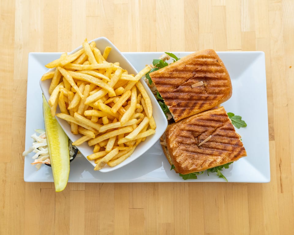 Grilled panini sandwich with fries, coleslaw, and a pickle spear on a white rectangular plate.