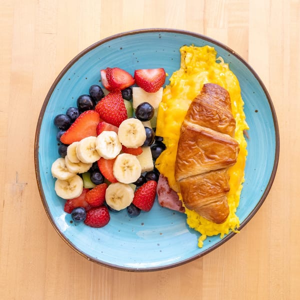 Breakfast plate with scrambled eggs, ham, croissant, and a fruit salad on a blue plate.