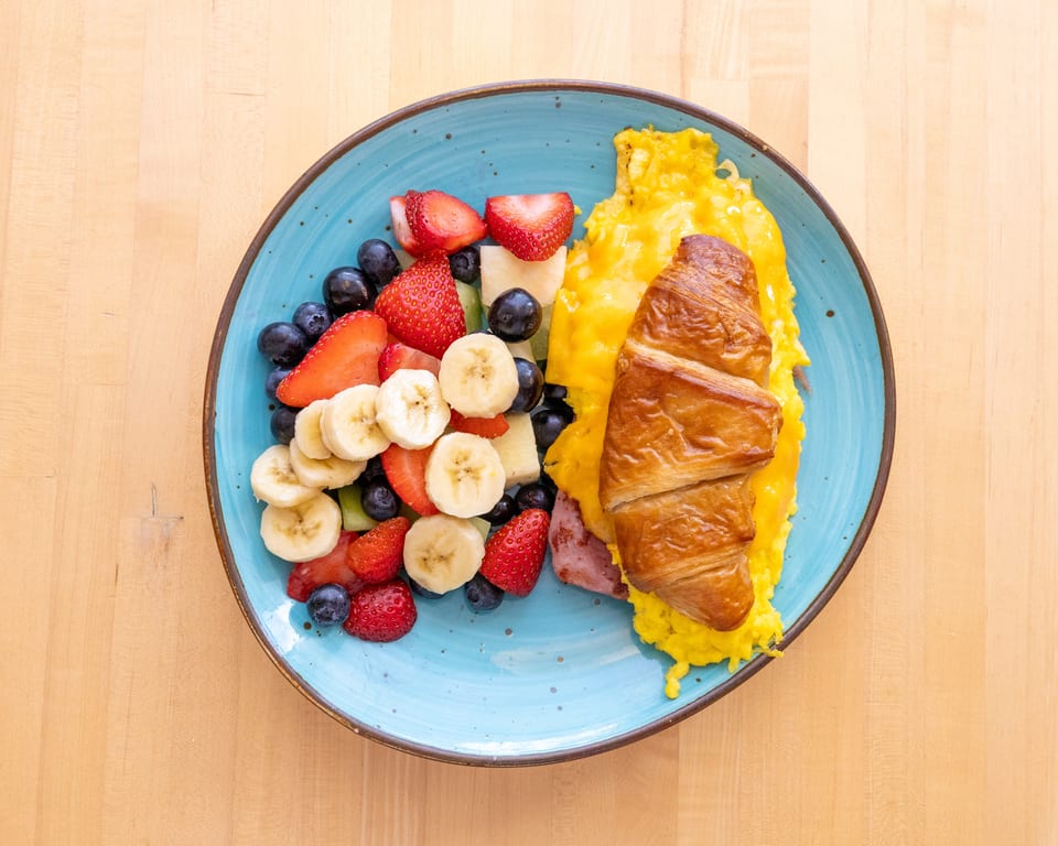 Breakfast plate with scrambled eggs, ham, croissant, and a fruit salad on a blue plate.
