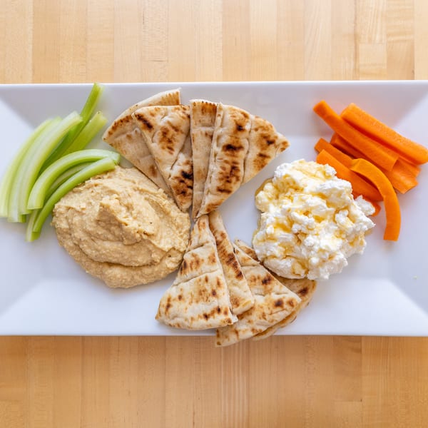 A white rectangular plate with hummus, pita bread, celery sticks, carrot sticks, and cottage cheese with honey.
