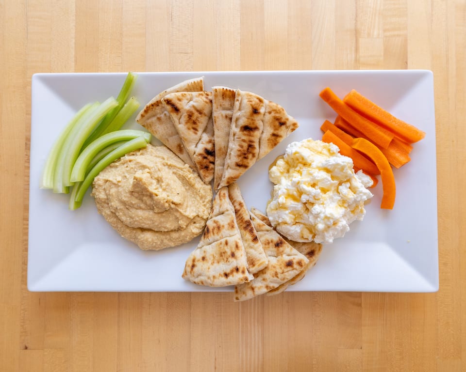 A white rectangular plate with hummus, pita bread, celery sticks, carrot sticks, and cottage cheese with honey.