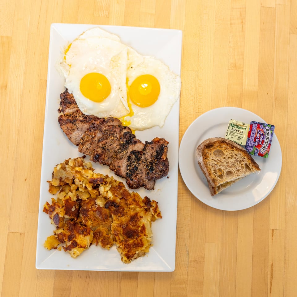 Steak and eggs breakfast with hash browns, toast, butter, and jelly on a wooden table.