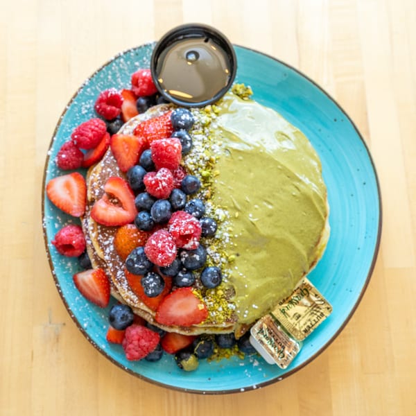 Pancakes with matcha cream, berries, powdered sugar, syrup, and butter on a blue plate.