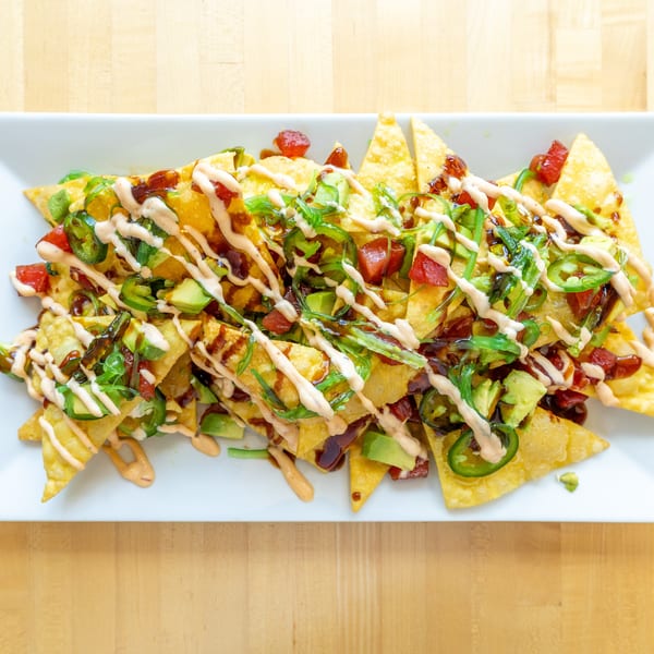 Crispy wonton nachos topped with tuna, avocado, jalapeños, seaweed salad, and sauces on a white plate.