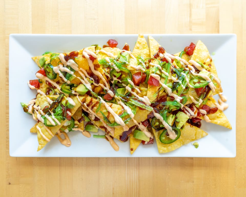 Crispy wonton nachos topped with tuna, avocado, jalapeños, seaweed salad, and sauces on a white plate.