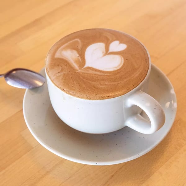 A white cup of latte with heart-shaped foam art sits on a wooden table with a spoon.