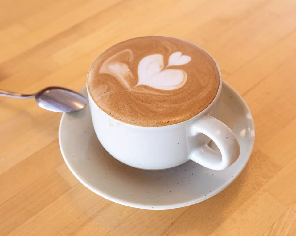 A white cup of latte with heart-shaped foam art sits on a wooden table with a spoon.