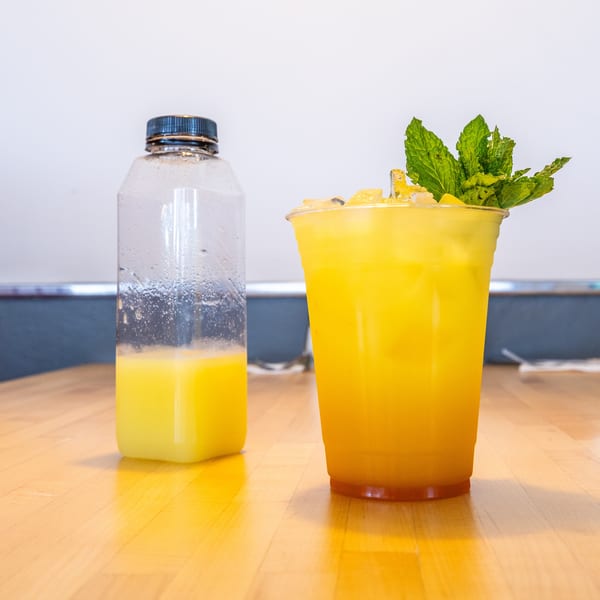 A plastic bottle with yellow liquid and a cup of iced yellow drink with mint on a wooden table.