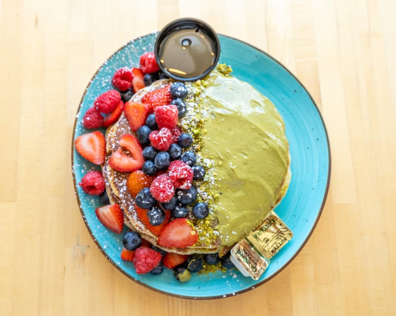Pancakes with matcha cream, fresh berries, powdered sugar, syrup, and butter on a blue plate.