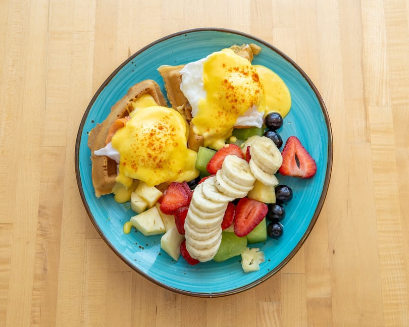Eggs Benedict on waffles with hollandaise sauce, served with a side of fresh fruit.