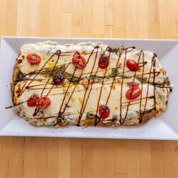 A rectangular flatbread pizza with melted cheese, pesto, cherry tomatoes, and balsamic glaze on a white plate.