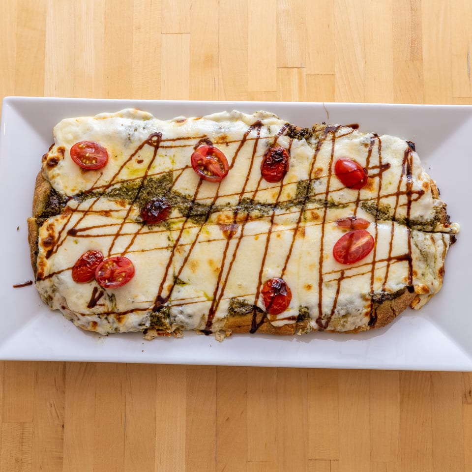 A rectangular flatbread pizza with melted cheese, pesto, cherry tomatoes, and balsamic glaze on a white plate.