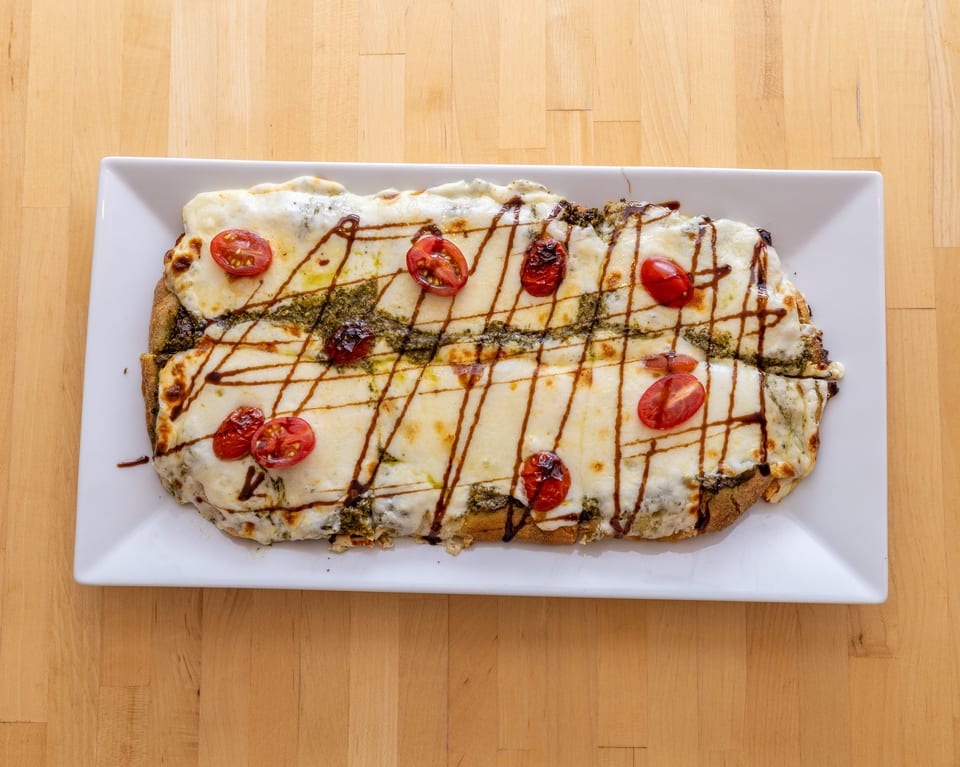 A rectangular flatbread pizza with melted cheese, pesto, cherry tomatoes, and balsamic glaze on a white plate.