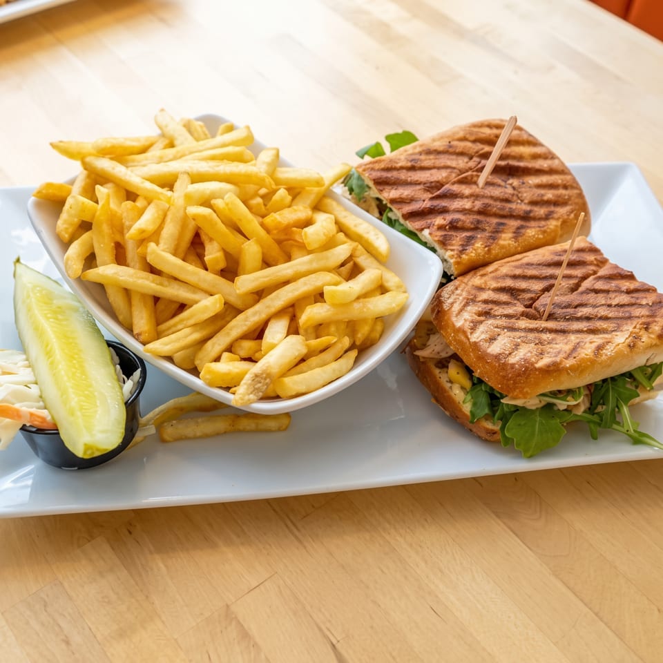 Grilled sandwiches with leafy greens and a bowl of golden fries served alongside pickles on a white platter.